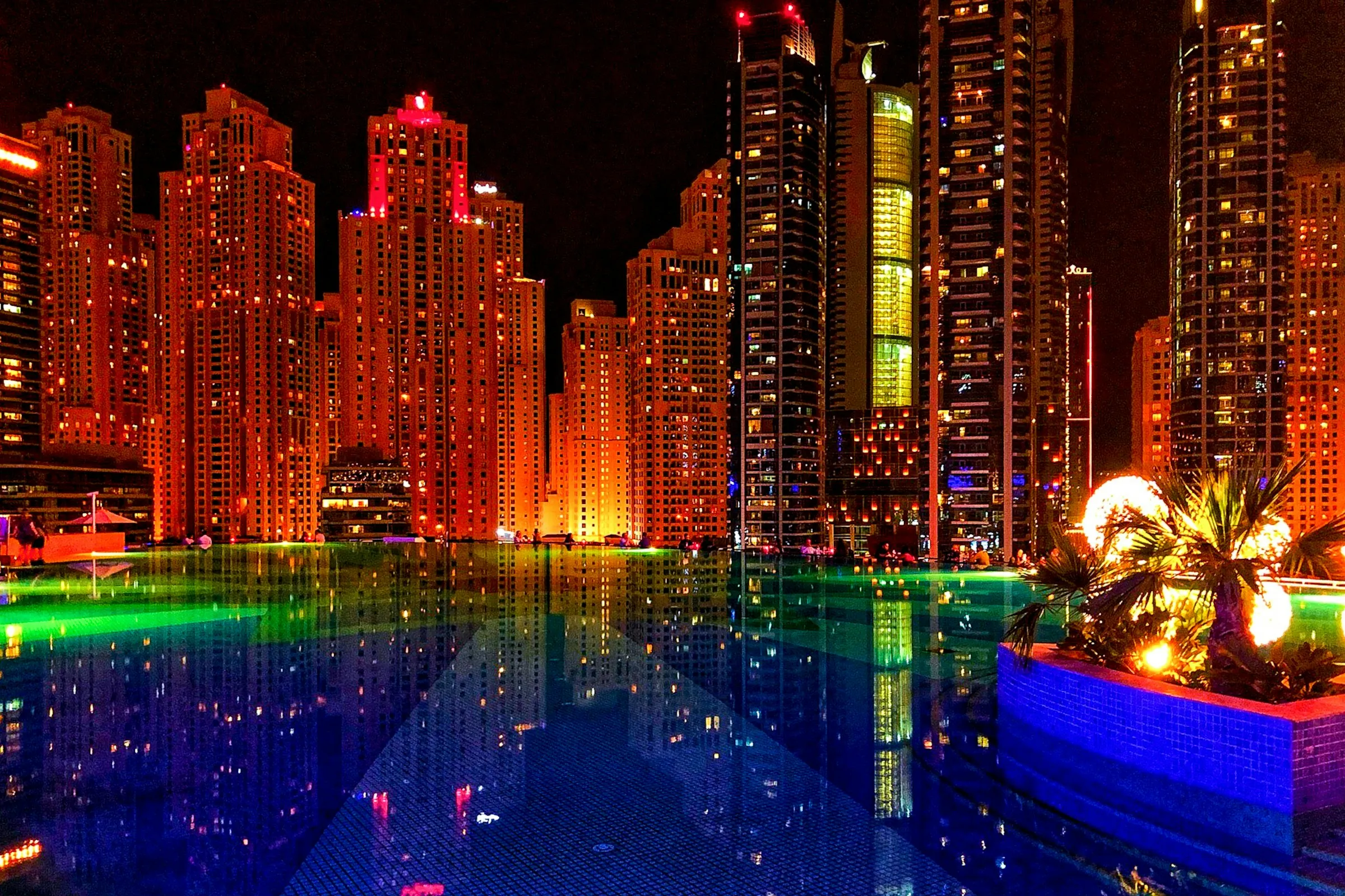 High-rise towers lit orange at night, reflected in a pool with coloured lighting