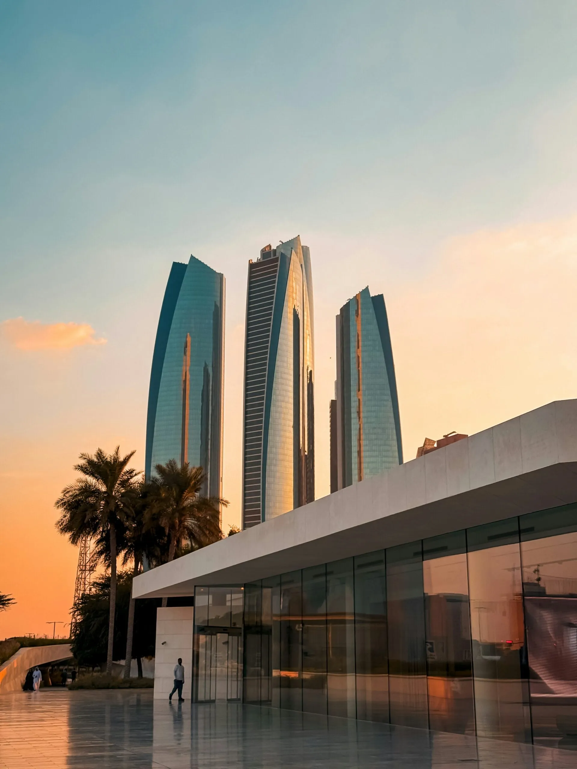 Etihad Towers at sunset behind a modern low-rise building and palm trees